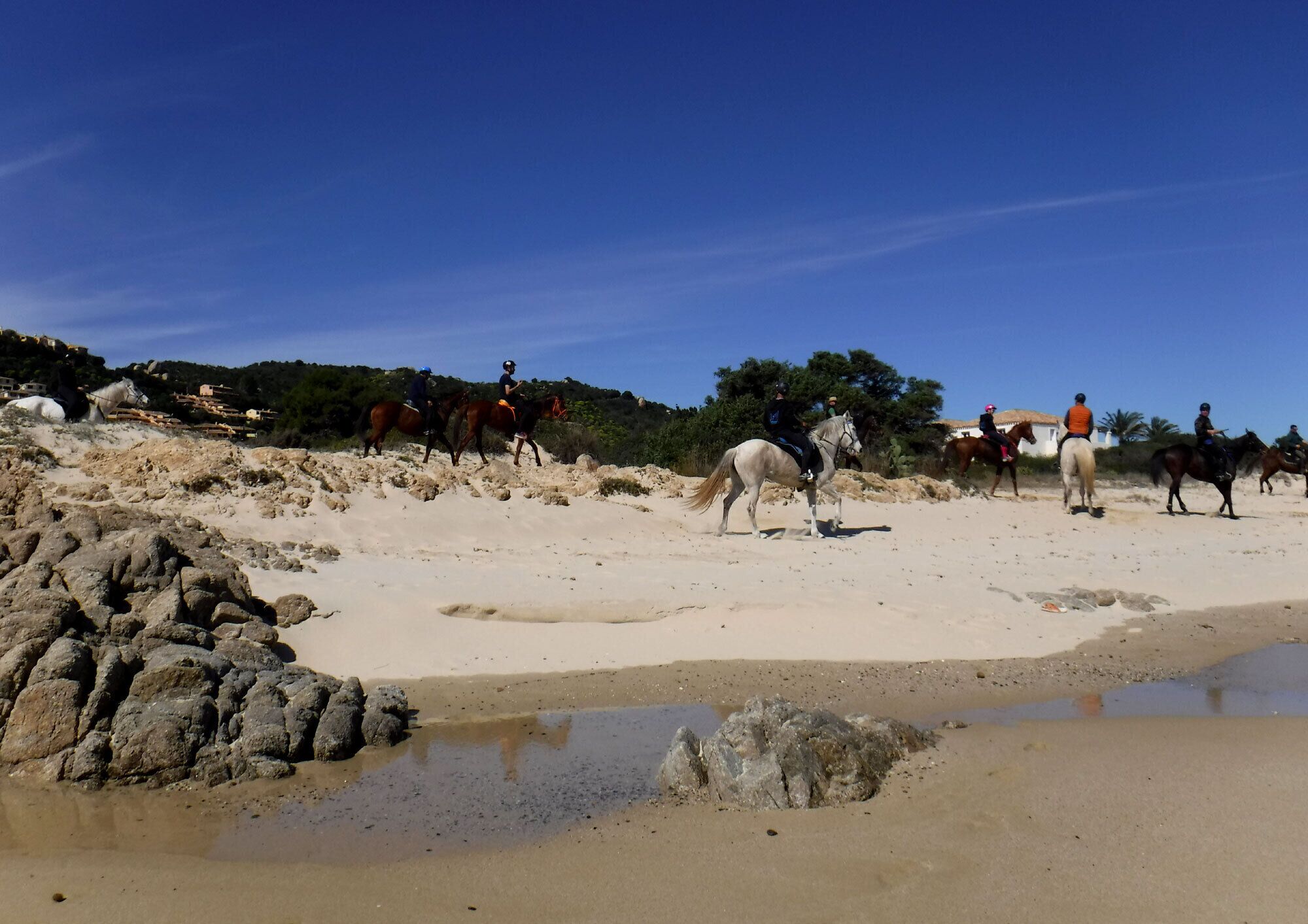 A beach with horses and riders, rocky terrain, and buildings in the background.