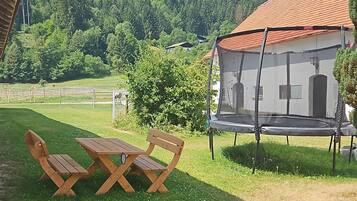 Een houten tafel met stoelen op een gazon, een trampoline, en een huis met een rood dak.
