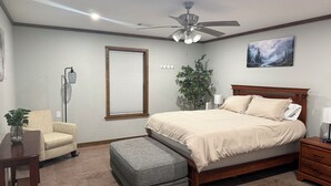 A bedroom with a bed, a nightstand, a chair, a floor lamp, a ceiling fan, a window with blinds, a plant, and a painting on the wall. - Wild Ride Retreat near Albert Pike Caddo River ATV trailer parking Pets allowed (Caddo Gap)