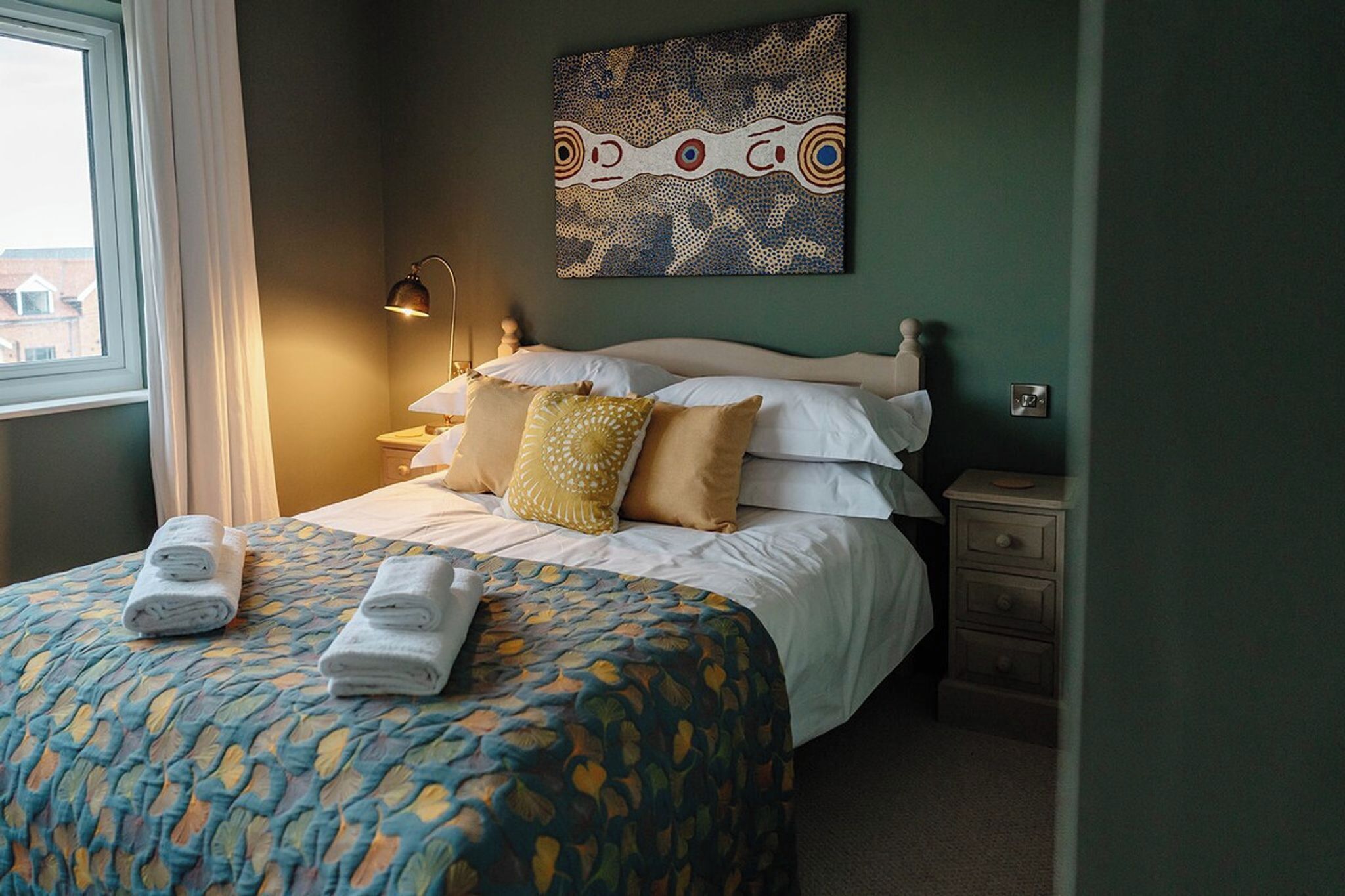 A neatly made bed with a patterned bedspread, a wall clock, and a painting above the bed.