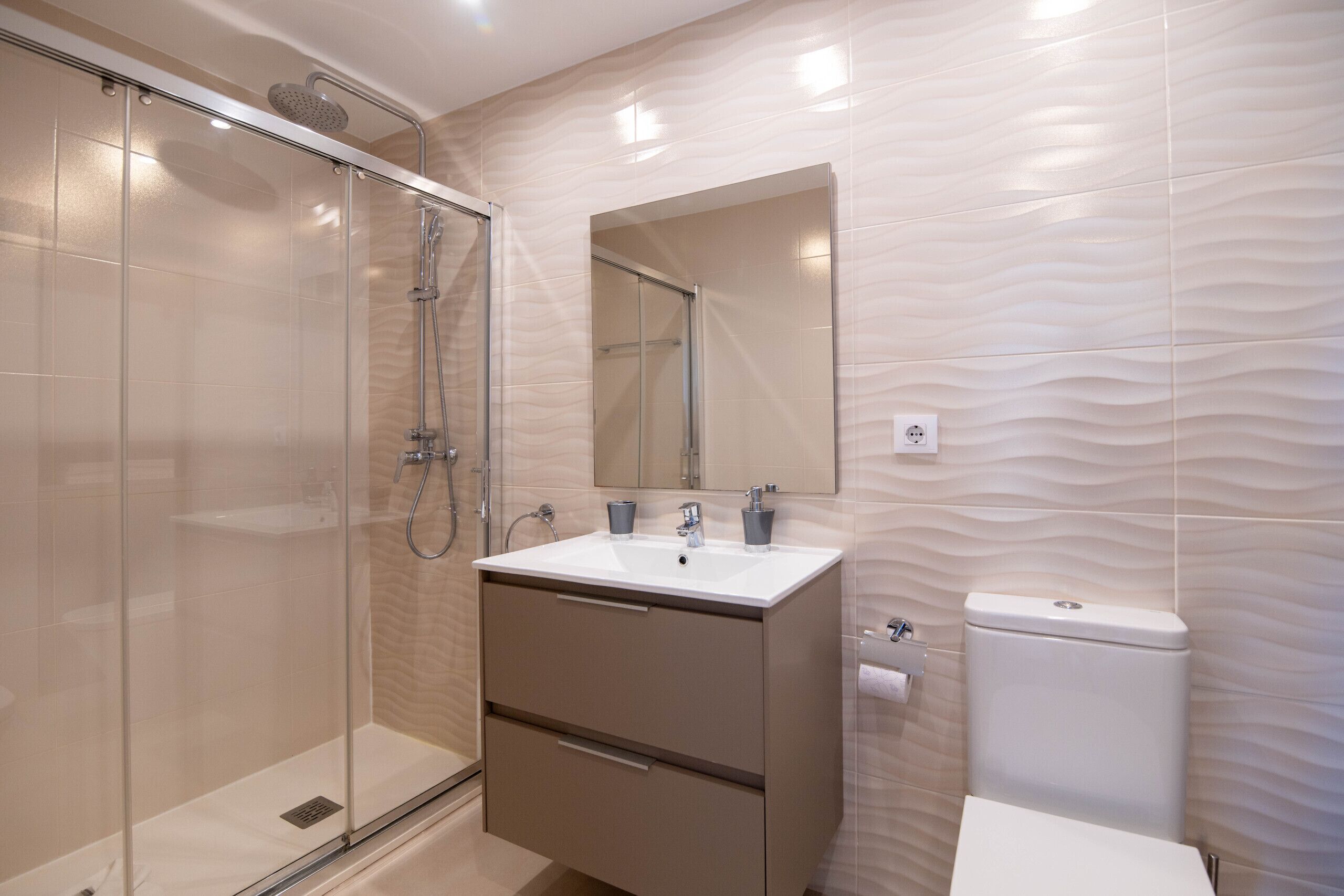 Un baño moderno con cabina de ducha de vidrio, tocador con lavamanos y espejo, y un inodoro.