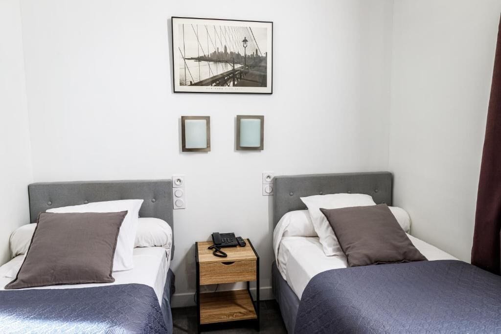 Een hotelkamer met twee bedden, een klein houten nachtkastje, een lijst met een afbeelding aan de muur en uitzicht op een stadslandschap.