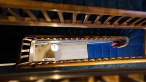 Una escalera de madera con barandilla de metal, una pared azul y el reflejo de una lámpara.