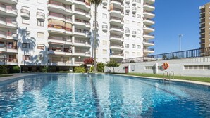 Uma piscina com superfície de água azul translúcida, cercada por um edifício de vários andares com varandas e um salva-vidas fixado na parede.