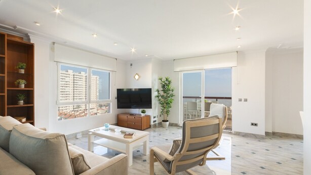 Uma sala de estar moderna com sofá, mesa de centro, televisão, estante de livros e varanda com vista para edifícios.