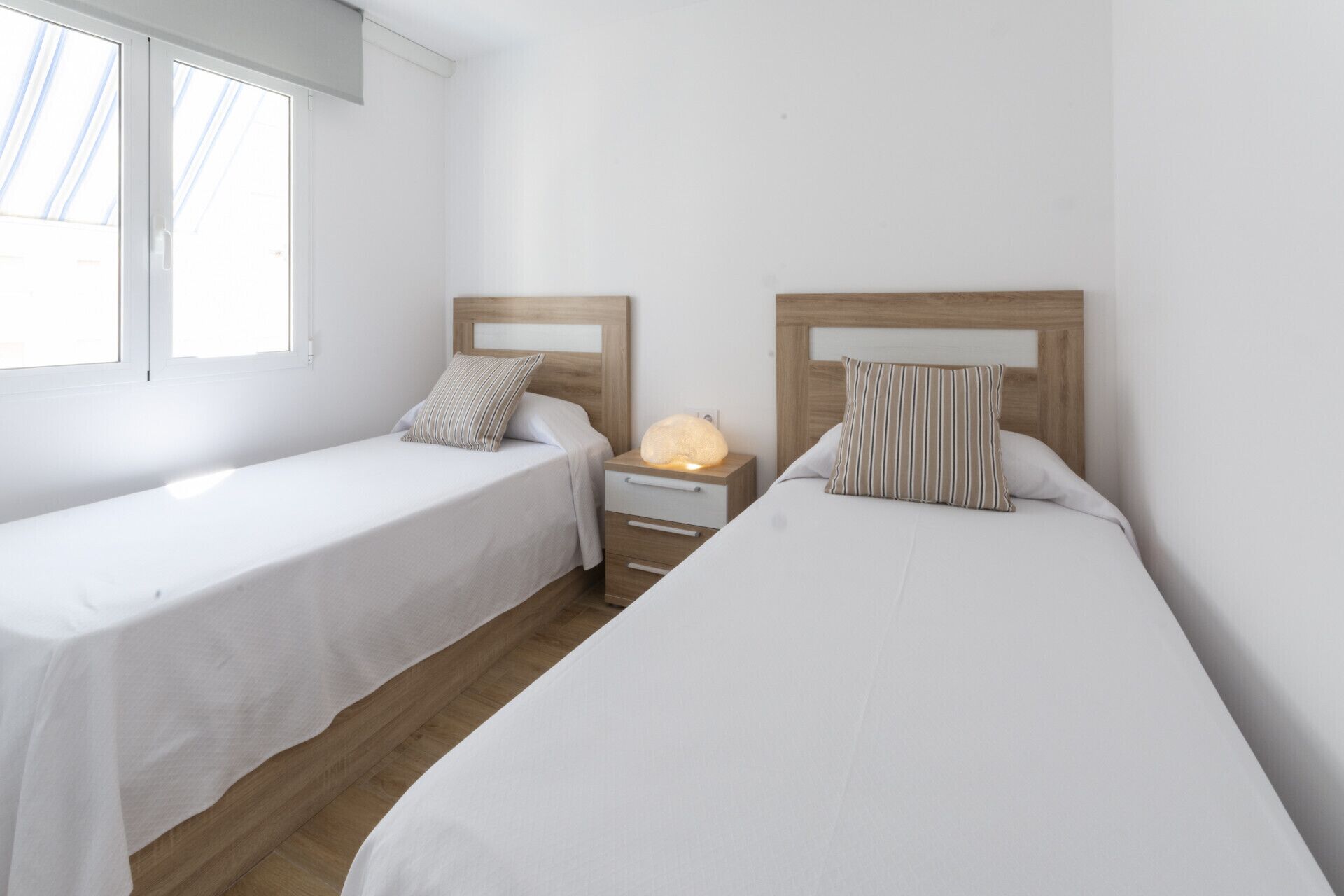 Una habitación con dos camas individuales, una mesita de noche con lámpara y una ventana.
