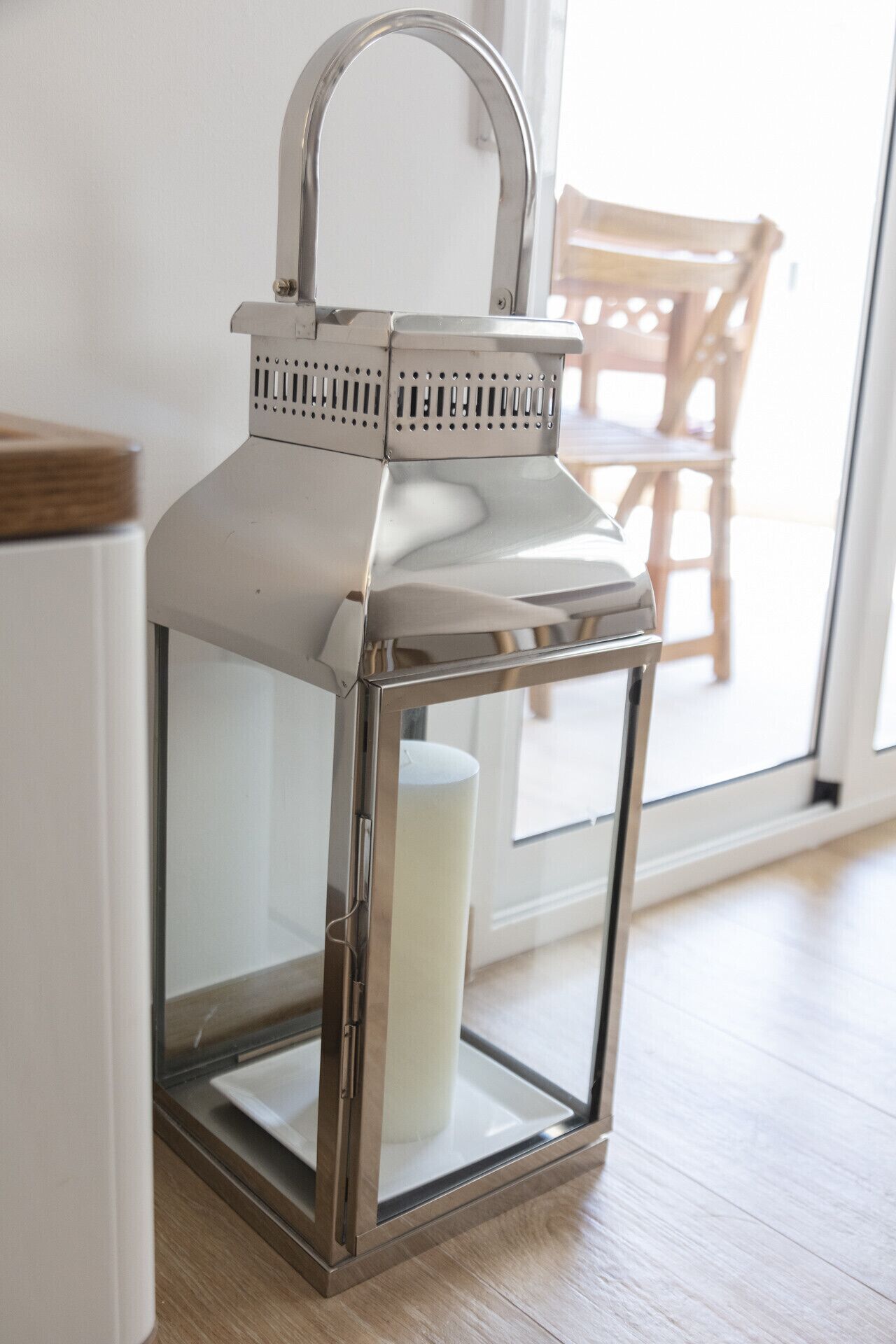 A metallic lantern with a handle and a cylindrical object inside, placed on a wooden floor against a white wall.