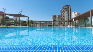 A swimming pool with tiled flooring, surrounded by wooden pergolas and benches, with tall residential buildings in the background. - Luxury vacations in Bermuda Apartments in front of the beach - ONLY FAMILIES (Playa de Gandía)