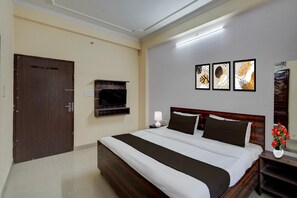 A hotel room with a bed, a television, a nightstand, a lamp, and framed artwork on the wall. - Deluxehotel Kartik Inn (Jaipur)