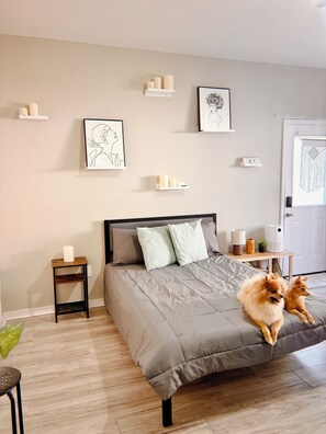 A bedroom with a bed, a nightstand, a small table, a door, and two dogs.