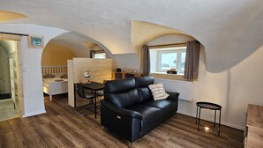 A room with a black leather sofa, a dining table, a small stool, and a window with curtains.