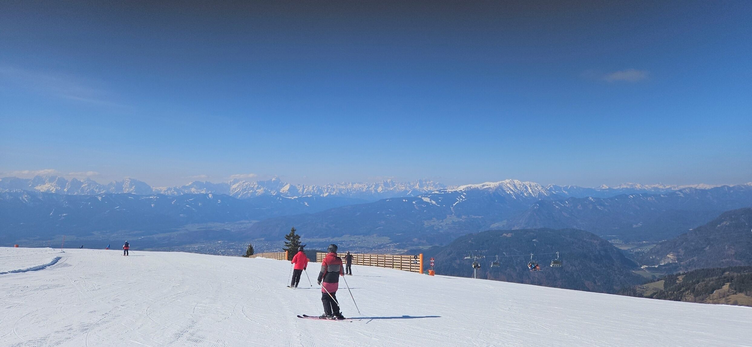 A snowy ski slope with skiers, a wooden fence, and a mountain backdrop.