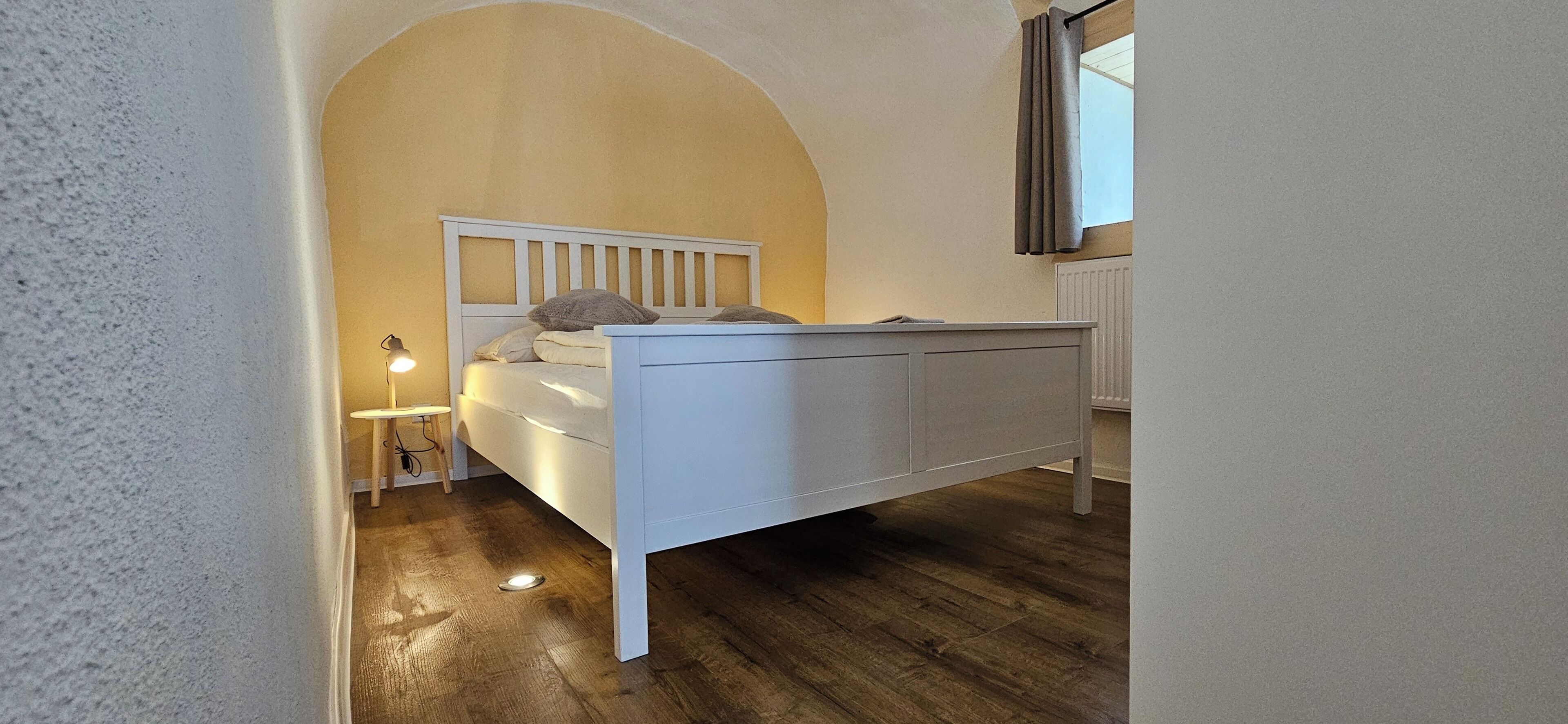 A bedroom with a white bed, wooden flooring, a small bedside table, and a window with curtains.