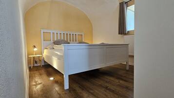 A bedroom with a white bed, wooden flooring, a small bedside table, and a window with curtains.
