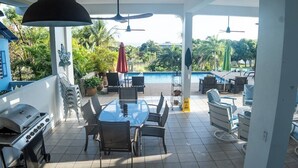 En udendørs terrasse med grill, spisebord, stole, parasoller og udsigt til en swimmingpool.