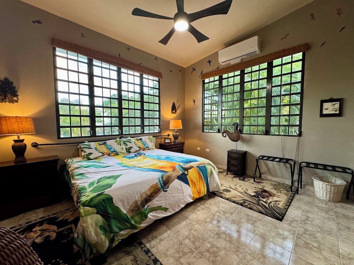 Une chambre à coucher comprenant un lit, deux fenêtres, un ventilateur de plafond, une table de chevet, une lampe, une chaise et un climatiseur fixé au mur.