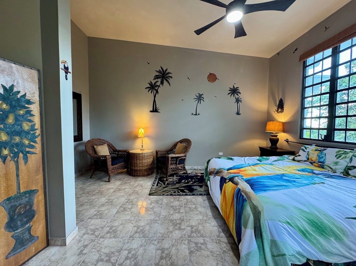 Un dormitorio con una cama, dos sillones de mimbre, una mesita, una lámpara y un mural en la pared.