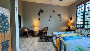 Un dormitorio con una cama, dos sillones de mimbre, una mesita, una lámpara y un mural en la pared.