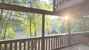 Ett trädäck med ett räcke, med utsikt över ett skogsområde, en sjö och en liten byggnad.