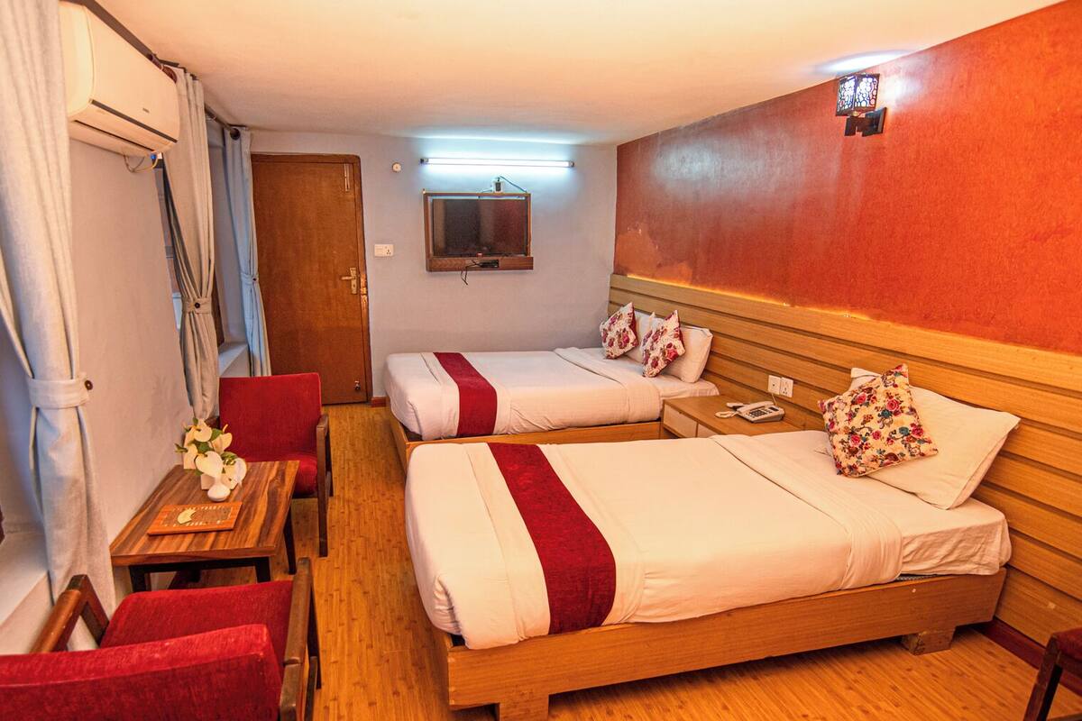 Camera d'albergo con due letti, una testiera in legno, un tavolino con un vaso di fiori, una sedia e un televisore a parete.