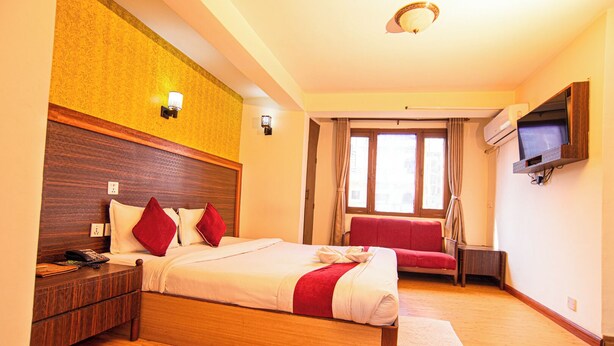 A hotel room with a wooden bed, a red sofa, a TV mounted on the wall, and a window with curtains. - Hotel Holiday Regency  (Kathmandu)