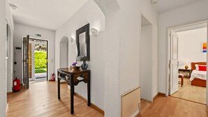 Un pasillo con piso de madera, una mesa con un florero, un espejo y una puerta que da a una habitación con cama y ventana.