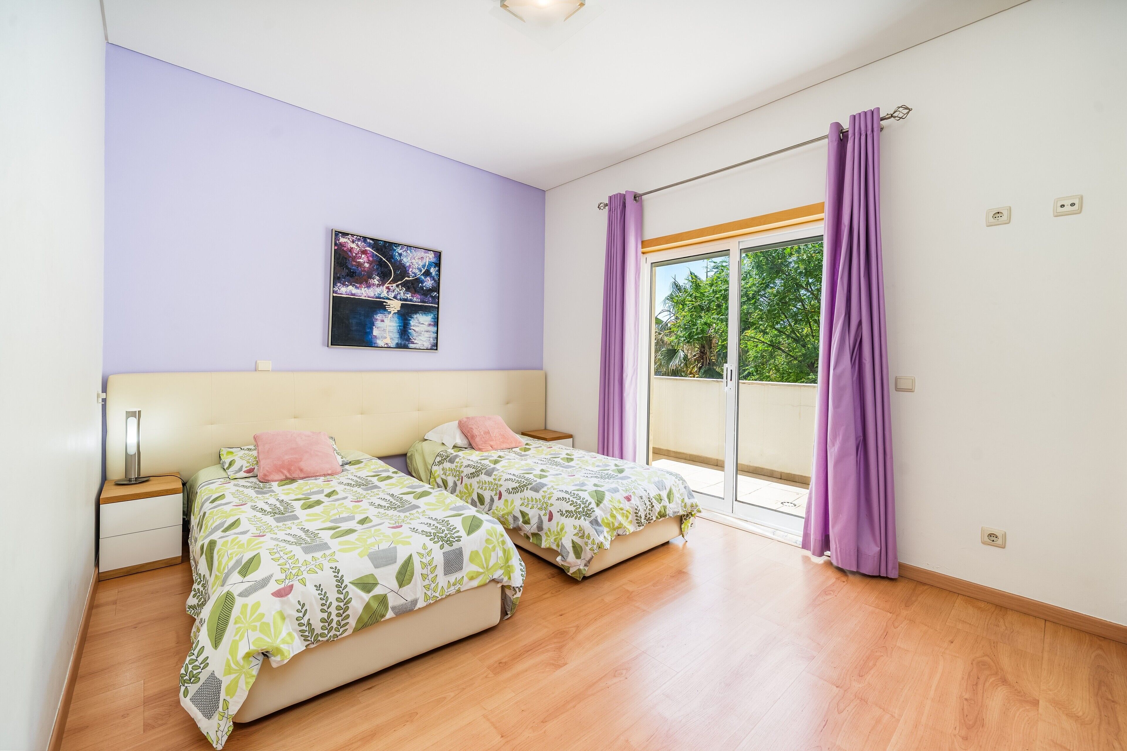 A bedroom with two beds, a nightstand, a wall-mounted lamp, a window with purple curtains, and a painting on the wall.