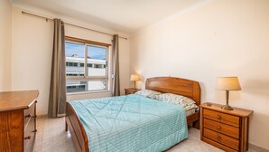 Een slaapkamer met een bed, een houten dressoir, een nachtkastje met een lamp en een raam met gordijnen.