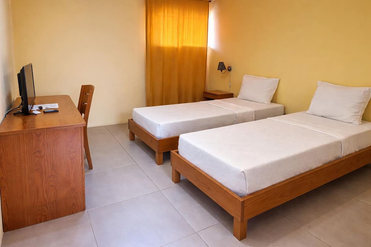 Una habitación con dos camas individuales, un escritorio, una silla, una lámpara y una cortina.