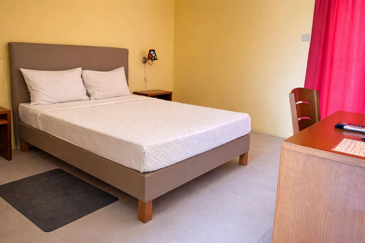 Una cama bien hecha con colchón y almohadas blancas, una mesita de noche de madera con una lámpara, un escritorio con una silla y una cortina roja.