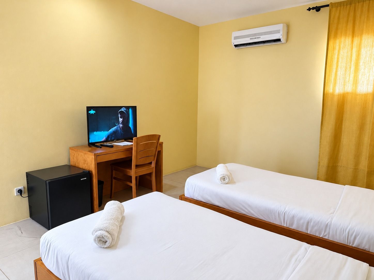 A hotel room with two beds, a desk with a computer, a television, and an air conditioning unit.