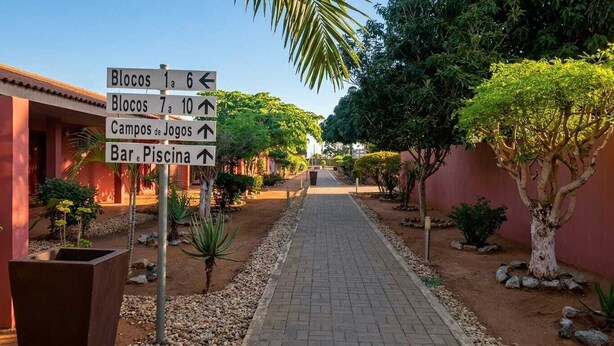 A pathway with directional signs pointing to various locations such as Blocos 1-6, Blocos 7-10, Campos de Jogos, and Bar/Piscina. - Viana Hotel (Viana)