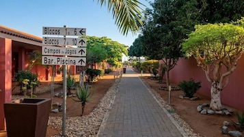 Un sendero con señales que indican diferentes lugares como Blocos 1-6, Blocos 7-10, Campos de Jogos y Bar/Piscina.