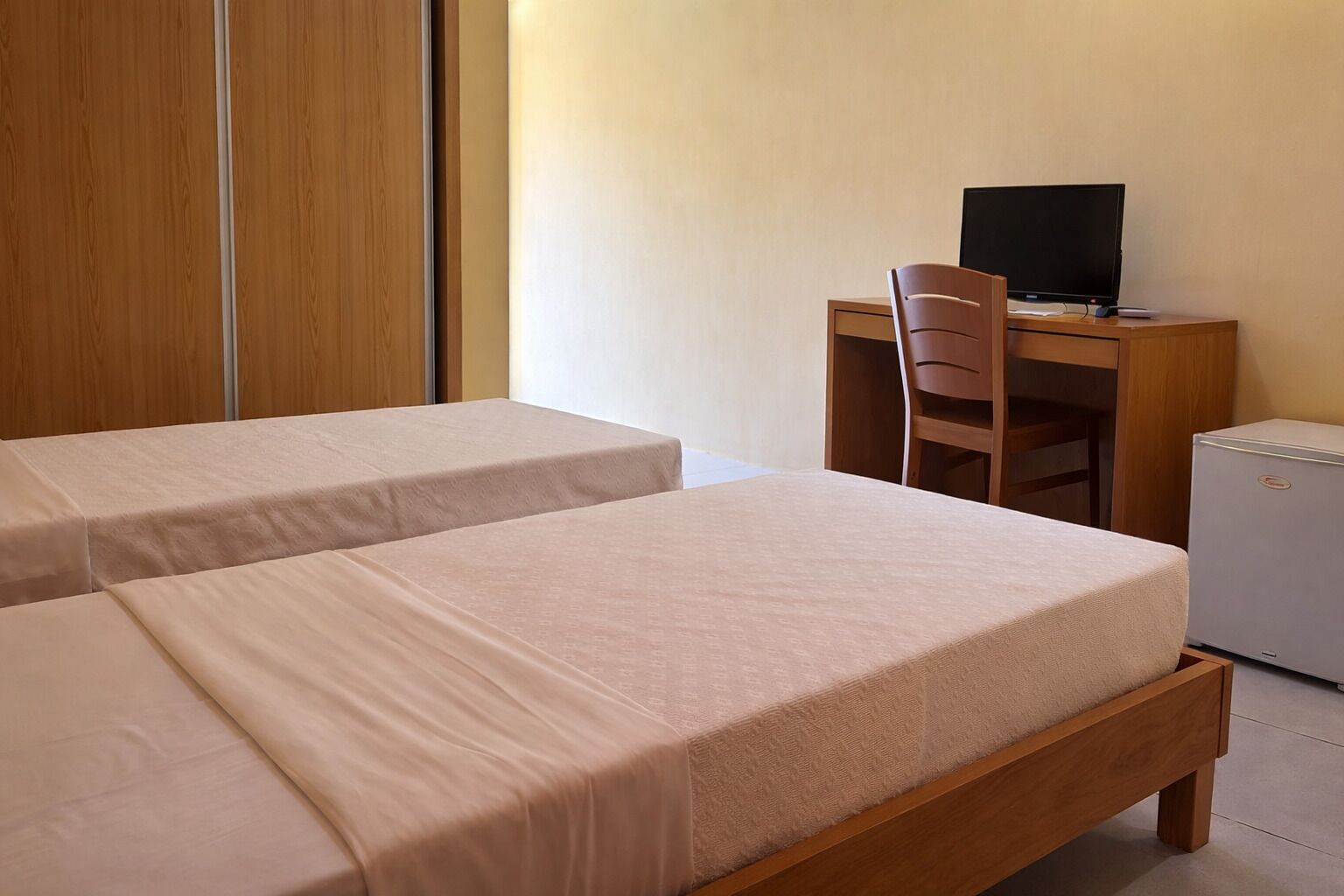A bedroom with two beds, a desk with a computer, a chair, and a small refrigerator.