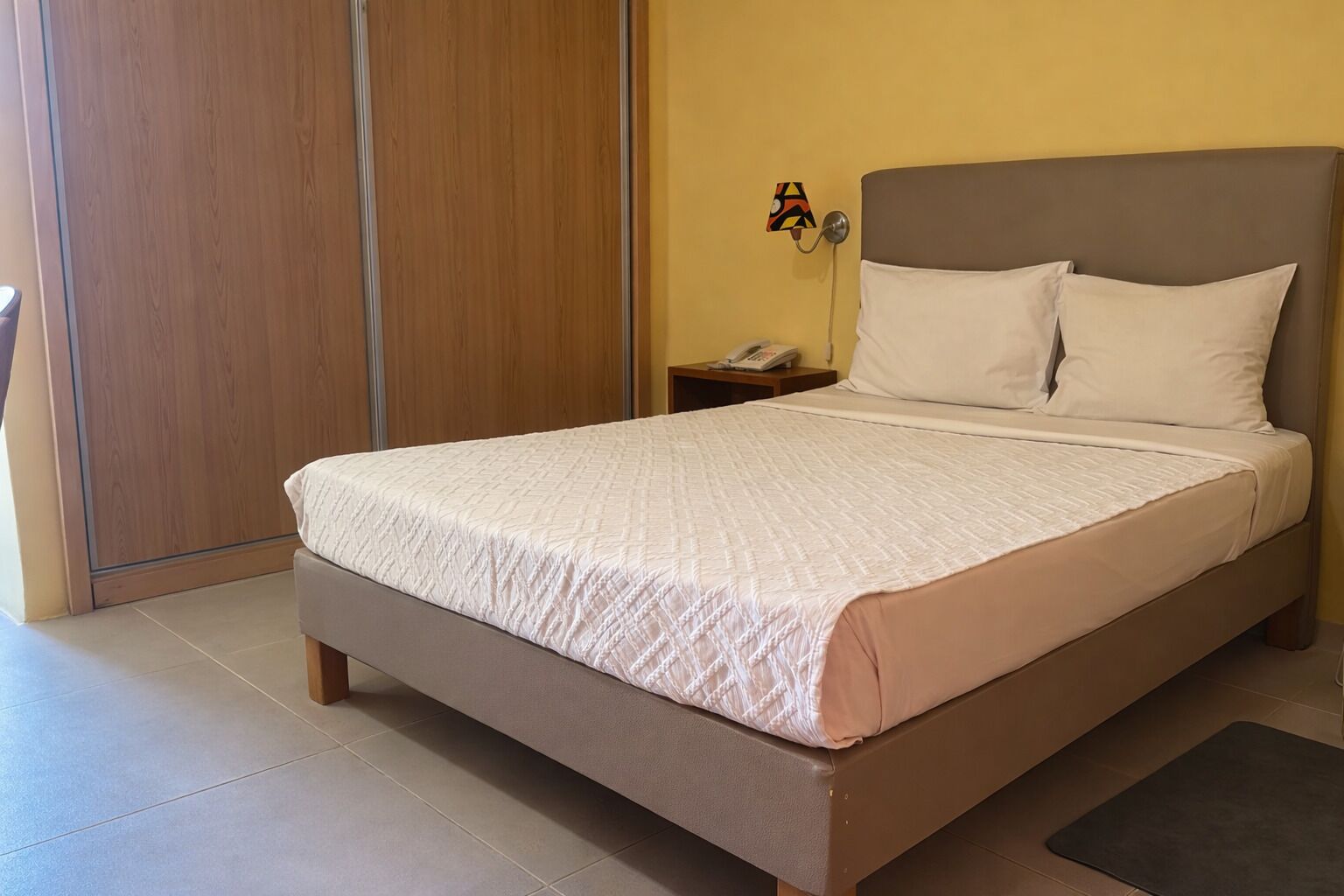 A single bed with a quilted white cover, a wooden headboard, a bedside table with a phone, and a wall-mounted lamp.