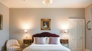 A bedroom with a large bed, two bedside tables, a chair, a door, and a chandelier. - Stuart Suite (Edinburgh)