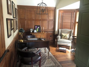A room with a wooden desk, leather chair, framed pictures, and a chandelier.