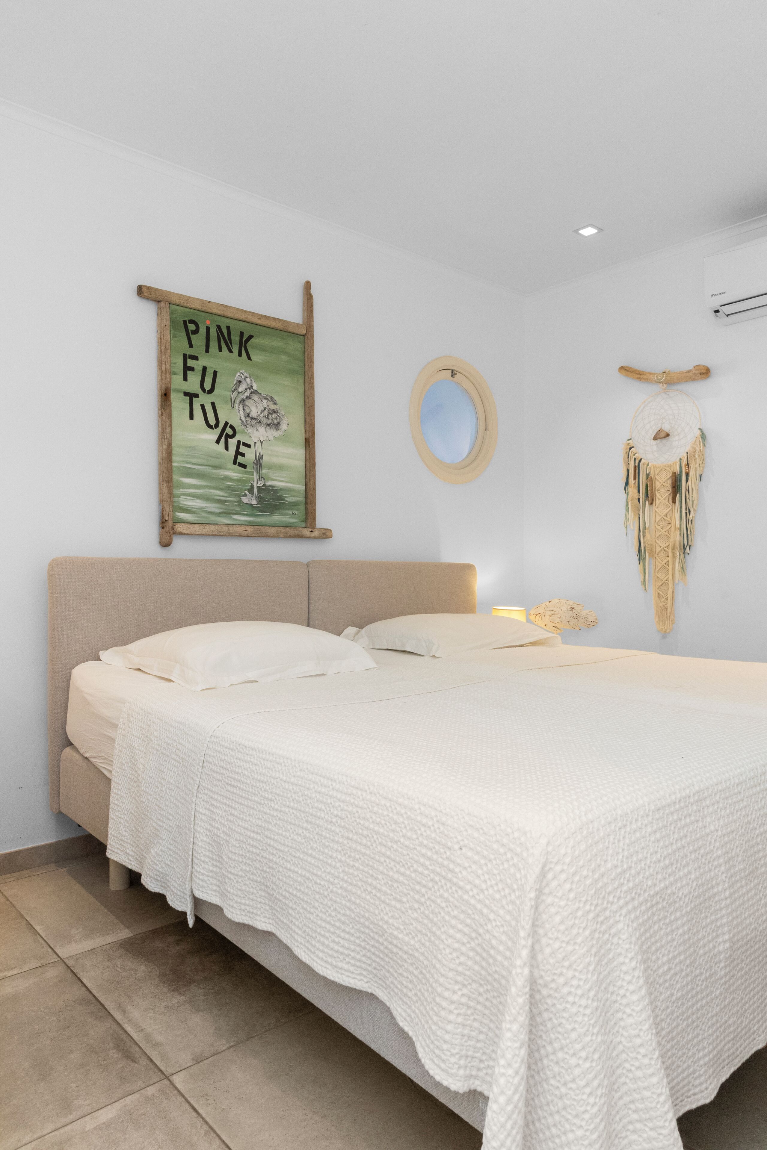 Una cama con cubrecama blanco, un espejo montado en la pared, un atrapasueños y un cartel en la pared.