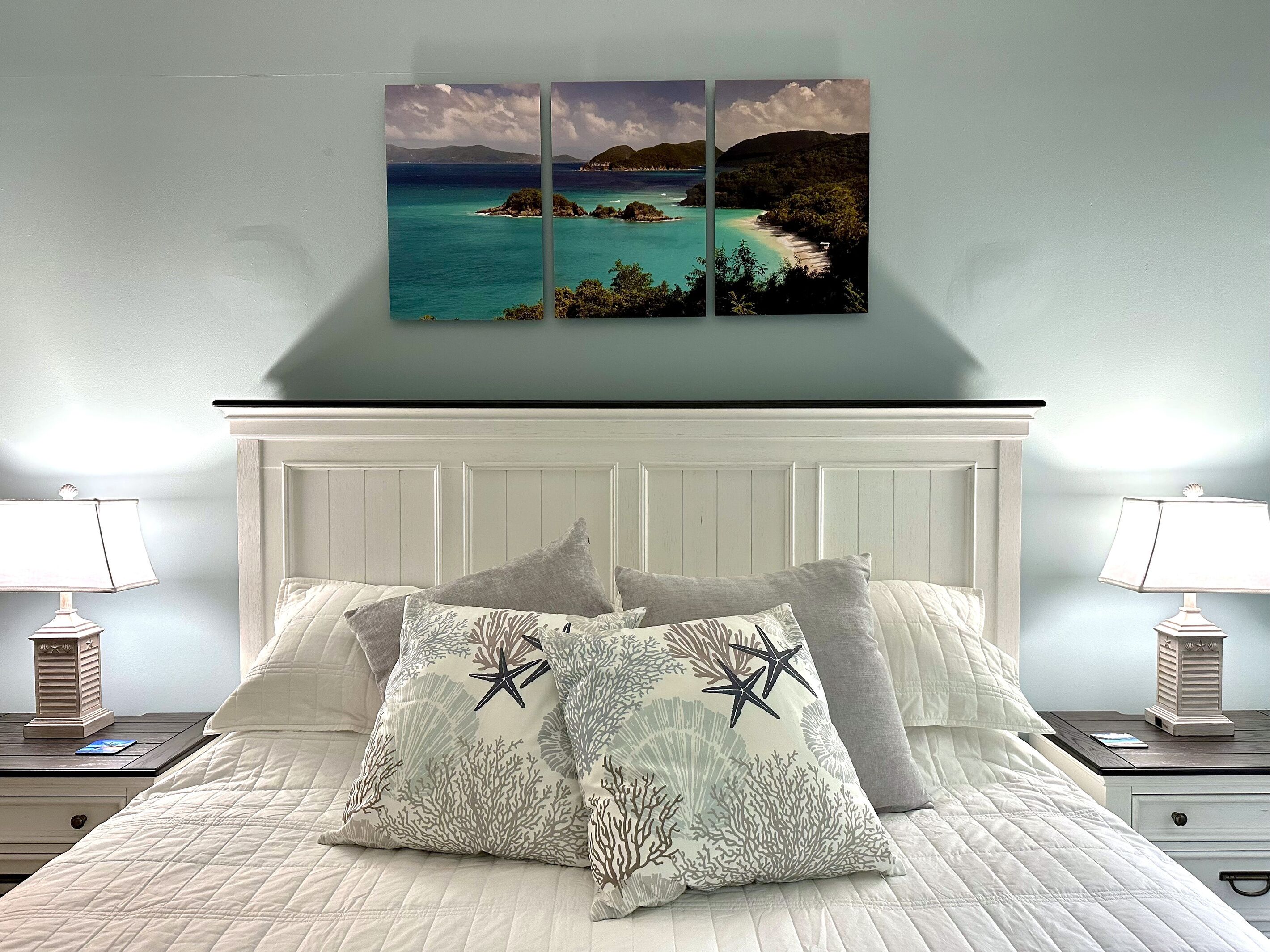 Un dormitorio con cabecera blanca, cama con ropa de cama blanca, dos mesitas de noche con lámparas y una obra de arte colgada en la pared que representa una escena costera.
