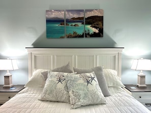 A bedroom with a white headboard, a bed with white bedding, two bedside tables with lamps, and a wall-mounted artwork depicting a coastal scene. - Fiesta @Serendip Vacation Condos (Cruz Bay)