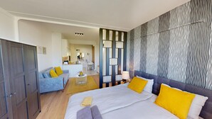 Quarto de hotel moderno com cama, sofá, escrivaninha e cozinha compacta.
