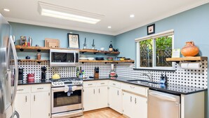 Una cocina moderna con armarios blancos, un refrigerador de acero inoxidable, un microondas, un horno y un lavavajillas. Hay repisas de madera sobre la encimera con diversos utensilios de cocina.
