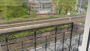 Een balkon met een metalen hekwerk met uitzicht op een spoorlijn en een bouwplaats met steigers.