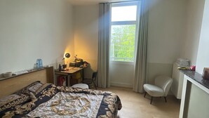 Een slaapkamer met een bed, bureau, stoel, ventilator en een raam met gordijnen.