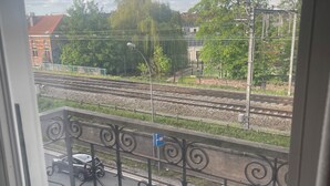 Uitzicht vanuit een raam op een balkon met een metalen hekwerk, een spoorlijn en een straat met een passerende auto.