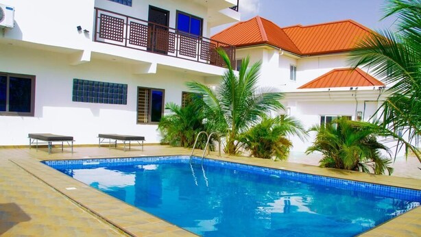 A swimming pool with a tiled deck, surrounded by palm trees and a white building with balconies. - Lawson Guest house  (Kweiman)