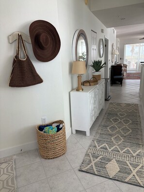 Un couloir avec un panier tressé, une console blanche, un tapis à motifs et un chapeau marron accroché au mur.