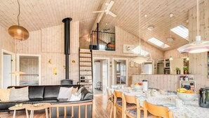 Un intérieur de chalet chaleureux avec un canapé, une table à manger, des chaises, un pouf noir, un espace en mezzanine et une cuisine.