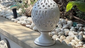 Un objeto decorativo blanco con diseños intrincados, sobre una superficie de piedra rodeada de rocas y follaje.