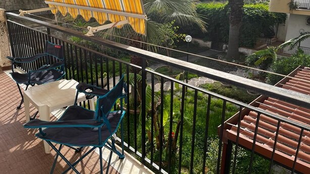 Un balcon avec des chaises, un auvent jaune à rayures et une vue sur des palmiers et un jardin.